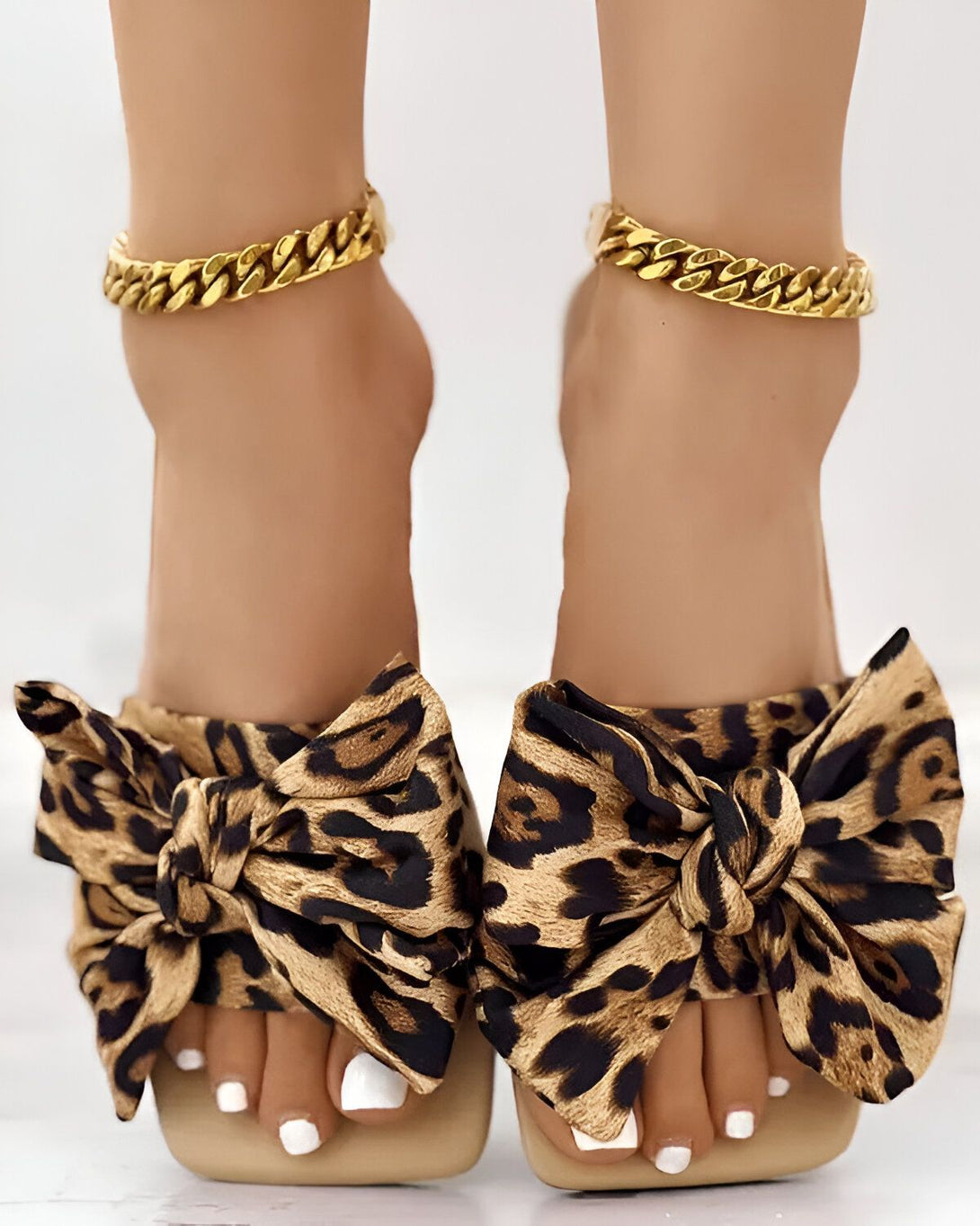 Une femme porte des sandales à talons hauts à bout ouvert. Les sandales sont en leopard et ont un nœud sur le devant. La femme a les ongles des pieds peints en blanc et porte des bracelets de cheville en or.
