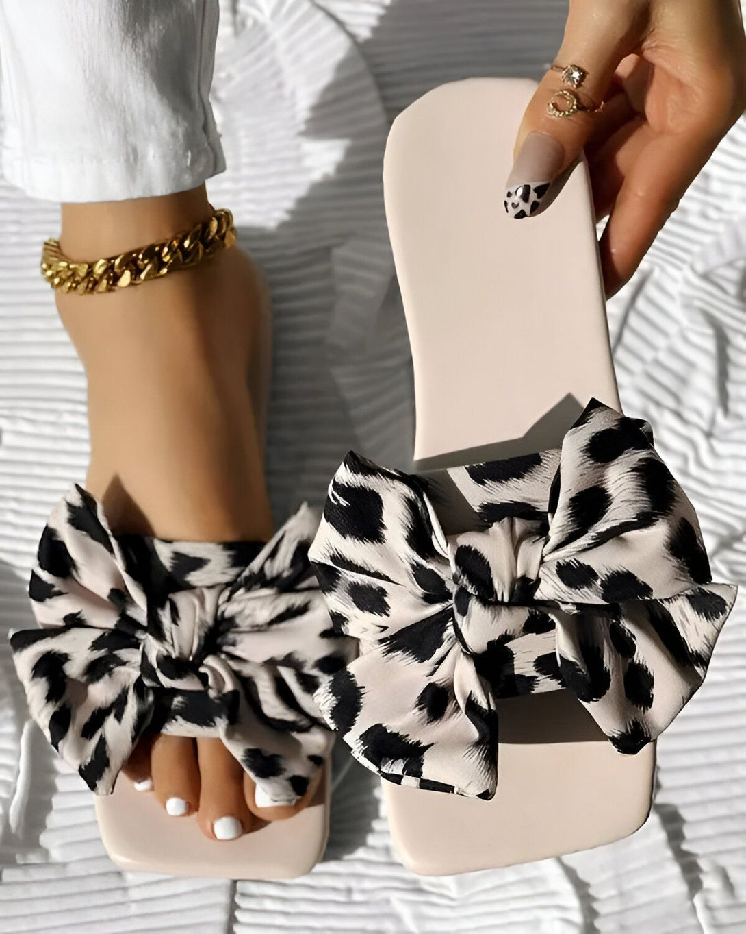 Une femme porte des sandales à talons hauts avec un nœud en satin léopard. Les sandales sont de couleur beige et le nœud est noir et marron. La femme a les ongles peints en noir et blanc. Elle porte également un bracelet de cheville en or.