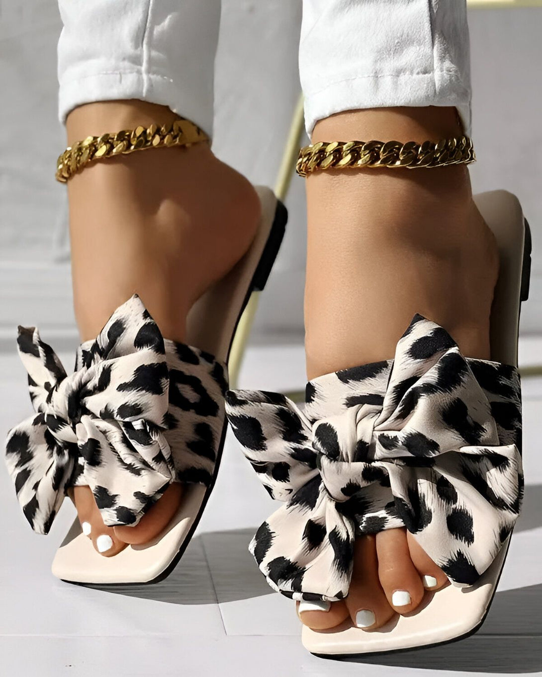 Une femme porte des sandales à talons hauts avec un nœud en tissu à imprimé léopard. Les sandales sont de couleur blanche et le nœud est noir et marron. La femme porte également un pantalon blanc et un bracelet de cheville doré.