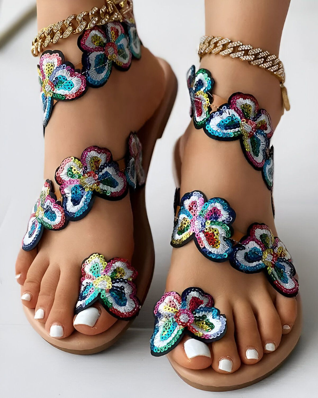Une paire de sandales plates à bout ouvert avec des fleurs en sequins multicolores. Les sandales sont portées par une femme avec une peau bronzée et des ongles blancs.