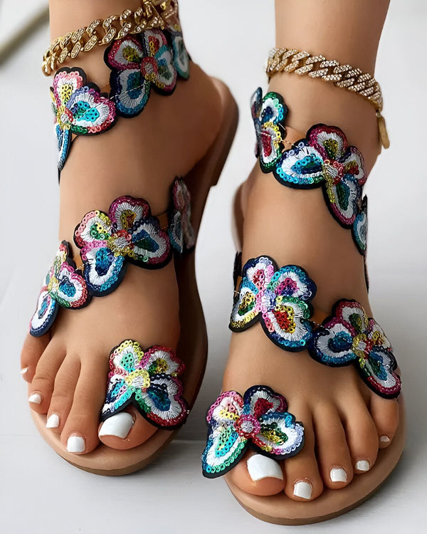 Une paire de sandales plates à bout ouvert avec des fleurs en sequins multicolores. Les sandales sont portées par une femme avec une peau bronzée et des ongles blancs.