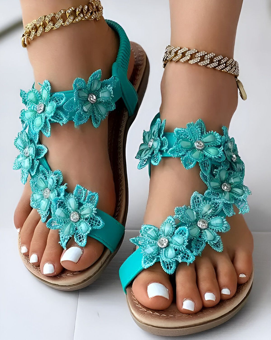 Une paire de sandales plates bleues avec des fleurs en tissu et des strass. Les sandales sont portées par une femme qui a les ongles des pieds peints en blanc.