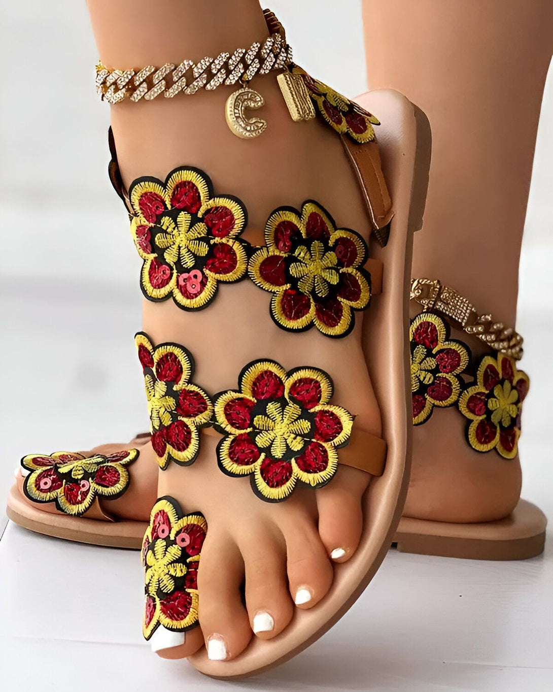 Une femme porte des sandales plates marron avec des fleurs rouges et jaunes brodées. Les sandales sont également dotées de brides à la cheville et de semelles antidérapantes.