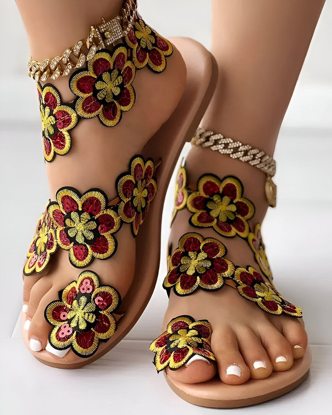 Une femme porte des sandales plates marron clair avec des fleurs rouges, jaunes et vertes brodées sur le dessus. Les sandales sont ouvertes au niveau des orteils et du talon et ont une bride qui s'enroule autour de la cheville. La femme porte également une chaîne dorée autour de la cheville.