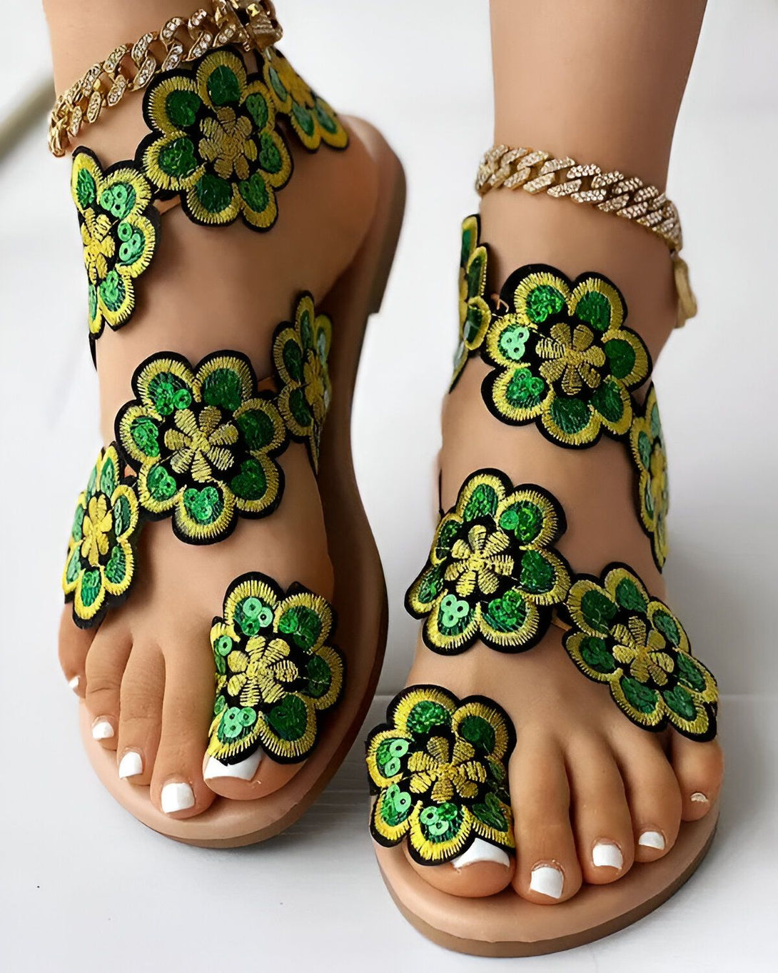Une paire de sandales plates vertes et jaunes à fleurs brodées. Les sandales sont portées par une femme qui a les ongles des pieds blancs et porte un bracelet de cheville en or.
