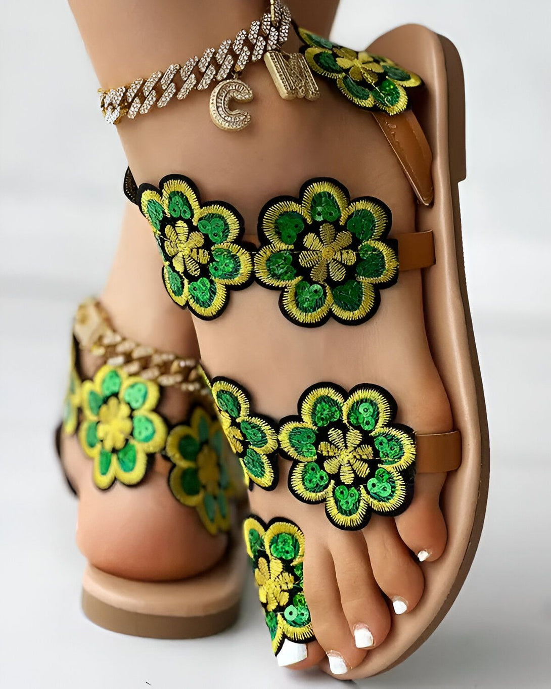 Une femme porte des sandales marron clair avec des fleurs vertes et jaunes brodées de sequins. Les ongles de ses pieds sont peints en blanc. Elle porte également une chaîne de cheville dorée avec un pendentif en forme de C.