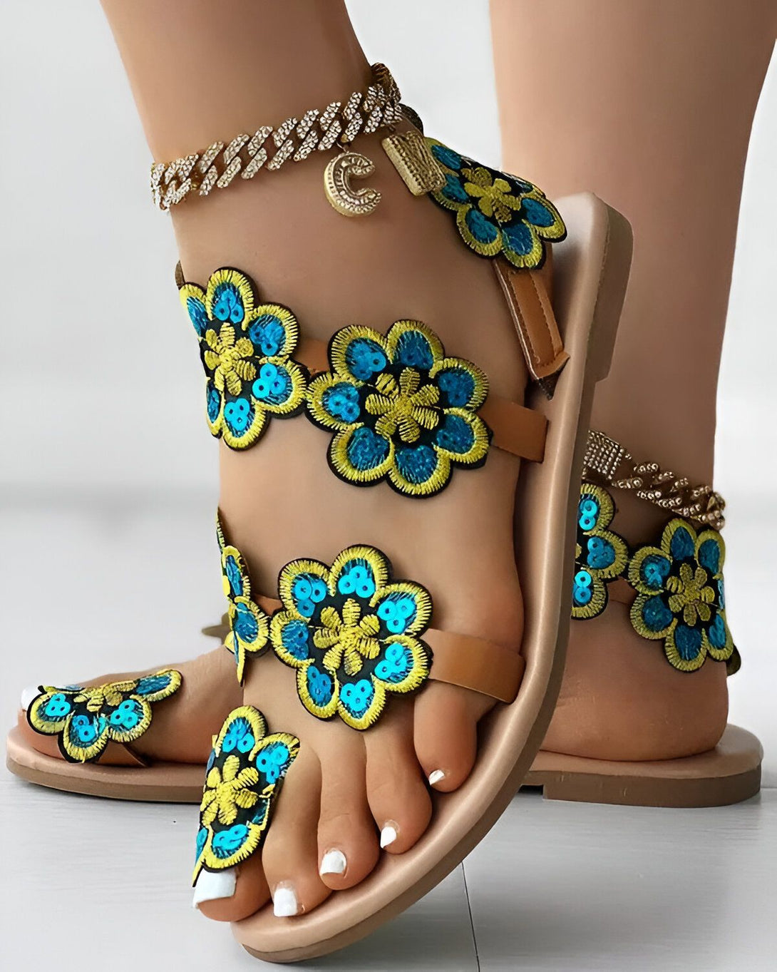 Une image de sandales plates marron avec des fleurs bleues et jaunes brodées. Les sandales sont portées par une femme qui a une chaîne de cheville en or avec un pendentif en forme de croissant de lune.