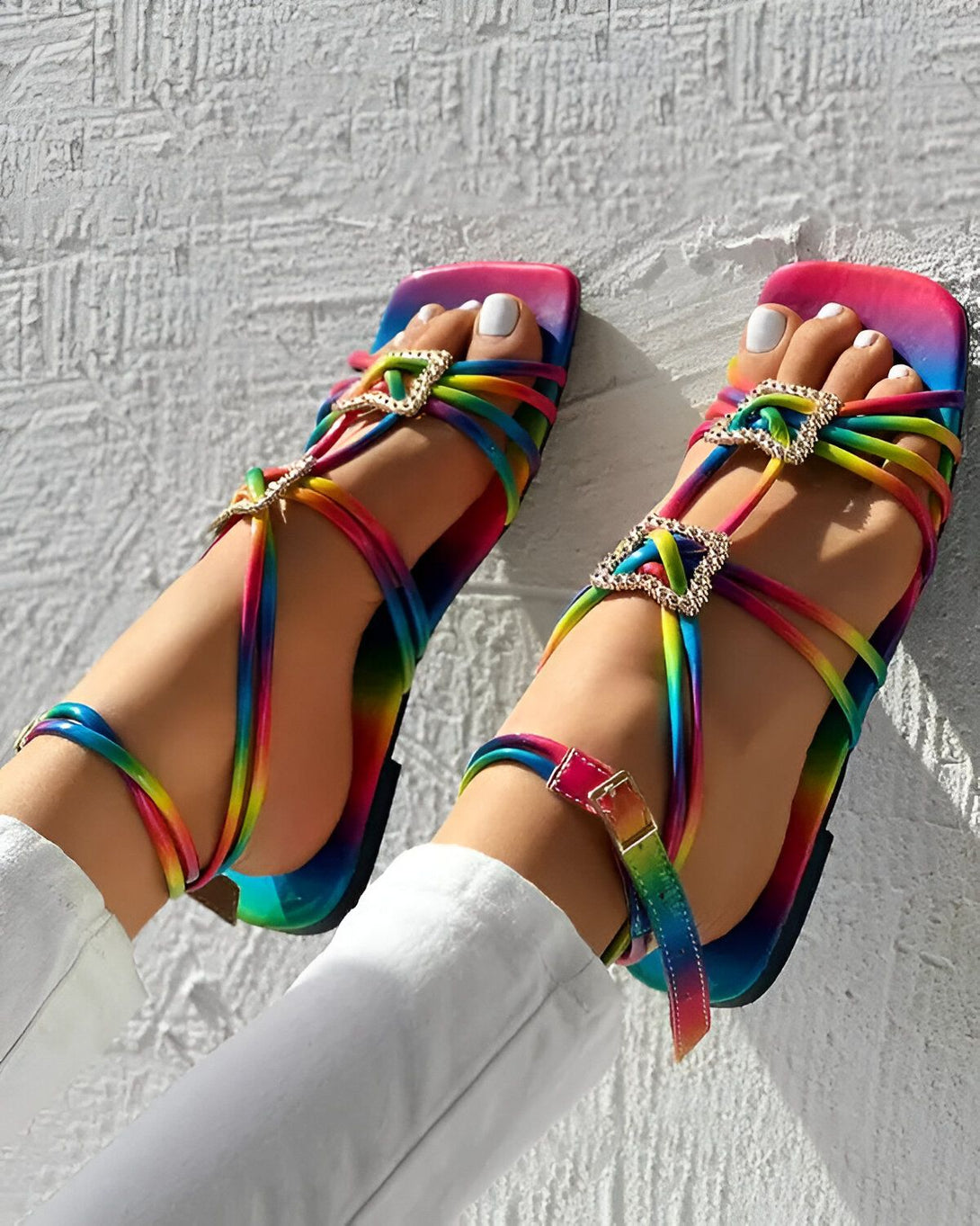 Une femme porte des sandales plates arc-en-ciel avec des brides croisées sur le pied et une boucle à la cheville.