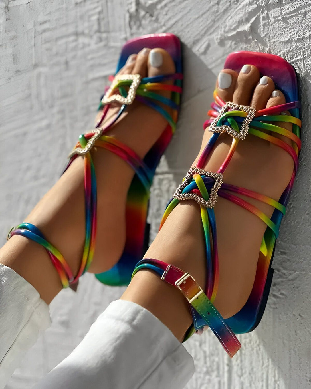Une femme porte des sandales arc-en-ciel à talons hauts. Les sandales sont attachées avec des boucles dorées. La femme porte un pantalon blanc et ses ongles sont peints en blanc.