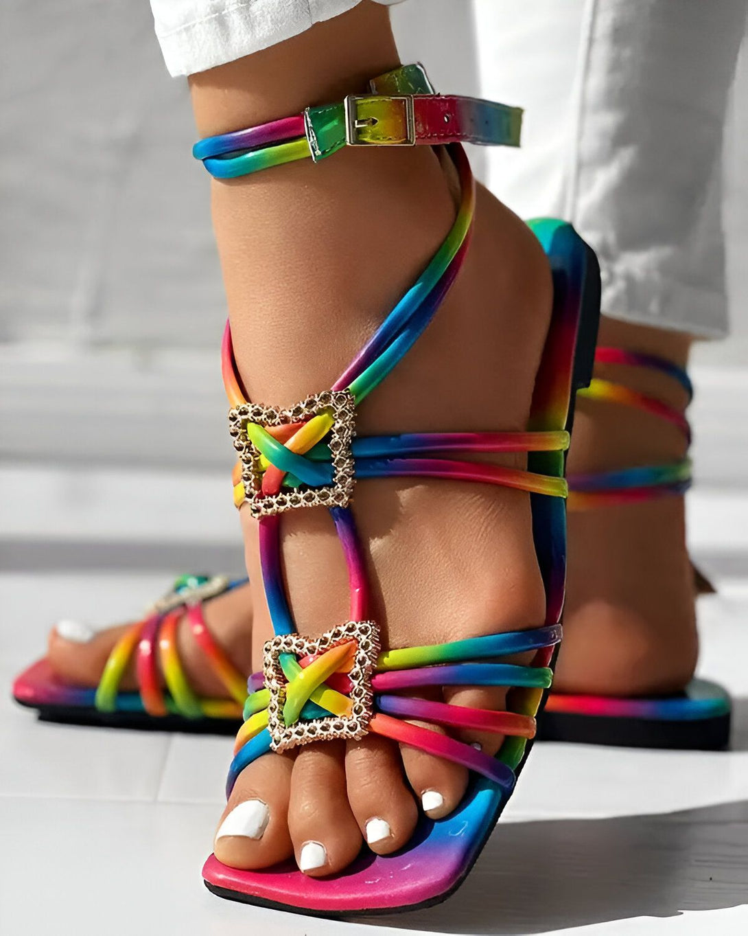 Une femme porte des sandales arc-en-ciel à talons hauts. Les sandales sont attachées à la cheville et ont des brides qui s'enroulent autour de la jambe. La femme porte un jean blanc et les ongles des pieds peints en blanc.