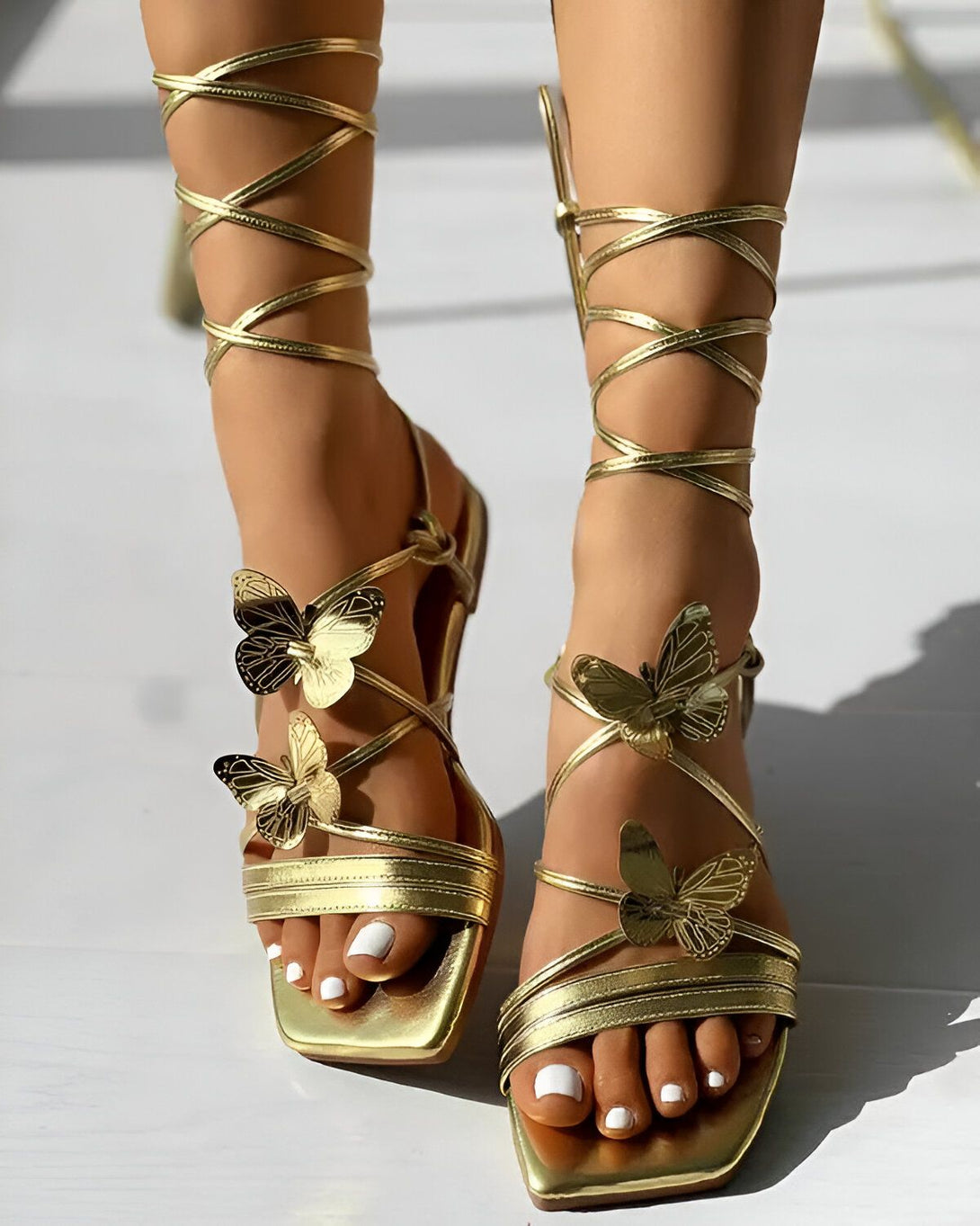 Une femme porte des sandales dorées à talons hauts. Les sandales sont ornées de papillons en métal doré. La femme a les ongles des pieds peints en blanc.