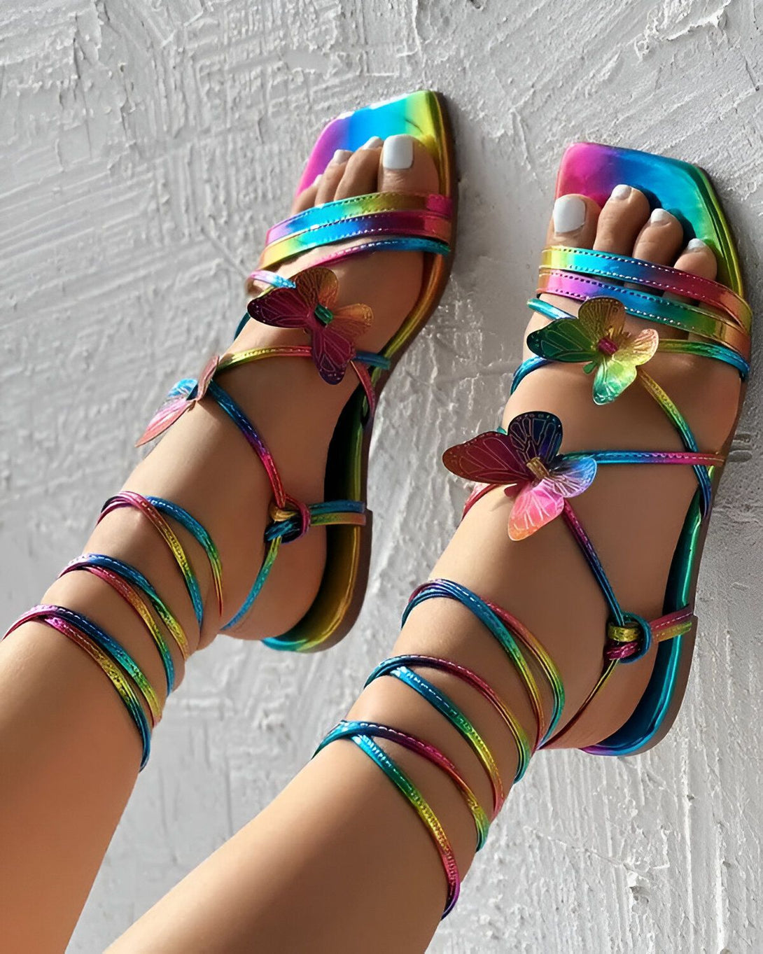 Une image de pieds avec des sandales colorées. Les sandales sont attachées aux chevilles avec des lacets arc-en-ciel. Les sandales ont des talons hauts et des bouts pointus. Les pieds sont vernis avec du vernis à ongles blanc.