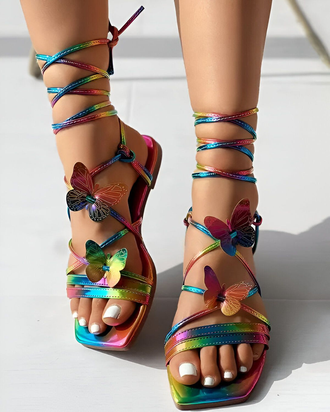 Une femme porte des sandales plates arc-en-ciel à lacets avec des papillons en 3D sur le dessus du pied.