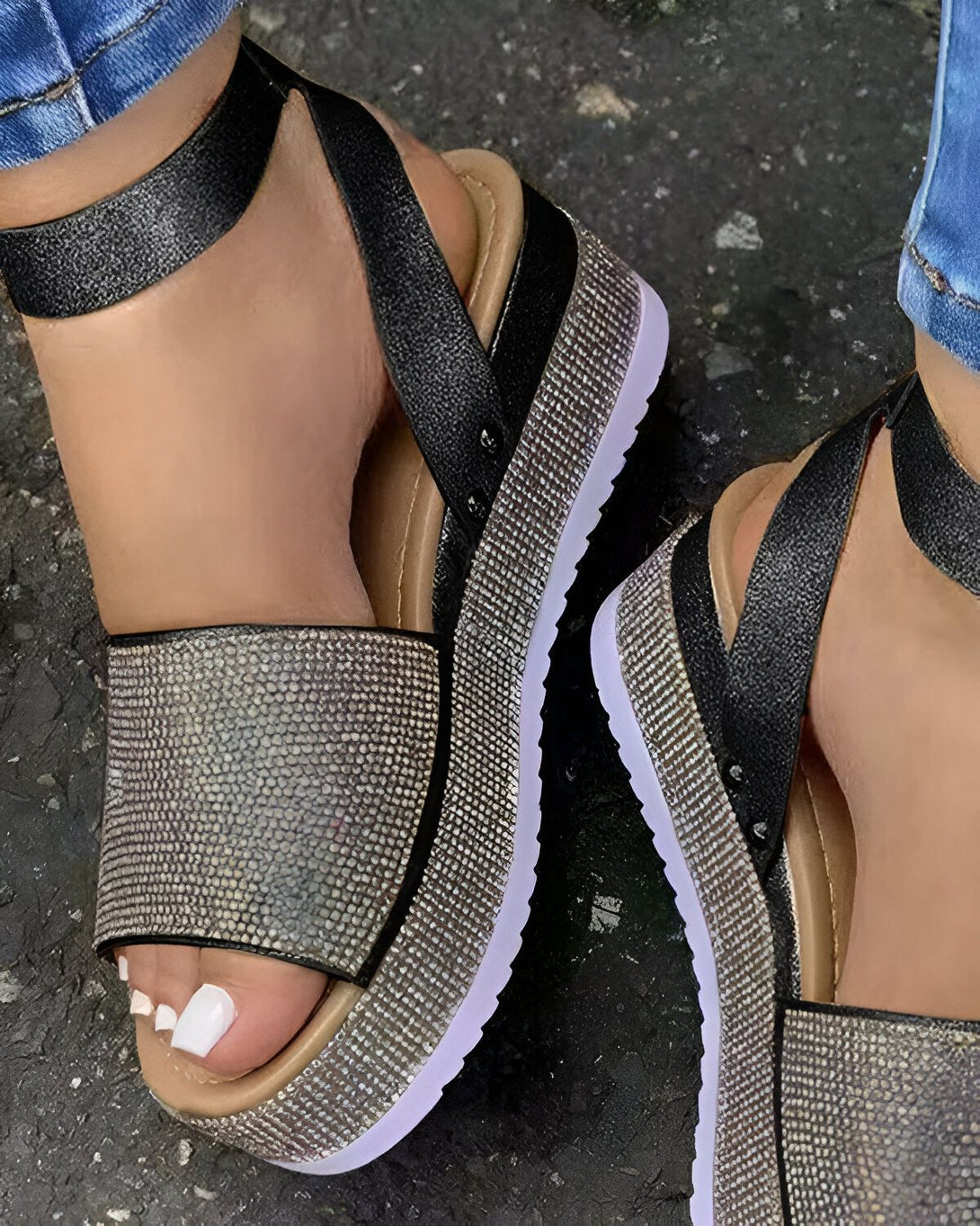 Une femme porte des sandales noires et argentées à plateforme. Les sandales sont en cuir et ont une bride à la cheville. La femme porte un jean bleu et ses ongles sont peints en blanc.