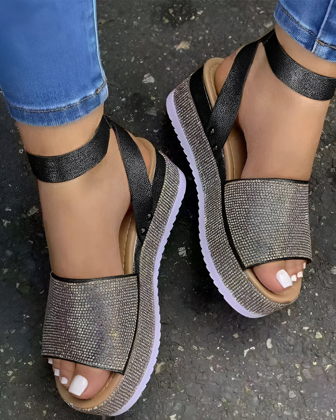 Une femme porte des sandales noires à plateforme avec des brides à scratch. Les sandales sont ornées de strass argentés. La femme porte un jean bleu et ses ongles sont peints en blanc.