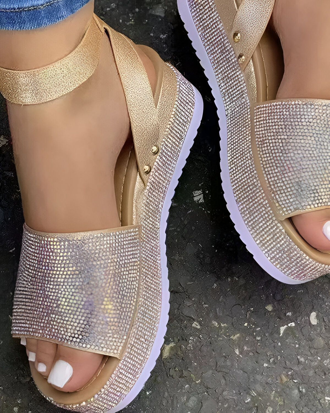 Une femme porte des sandales à plateforme dorées. Les sandales sont recouvertes de strass et ont une bride à la cheville. La femme porte un jean bleu et les ongles des pieds blancs.