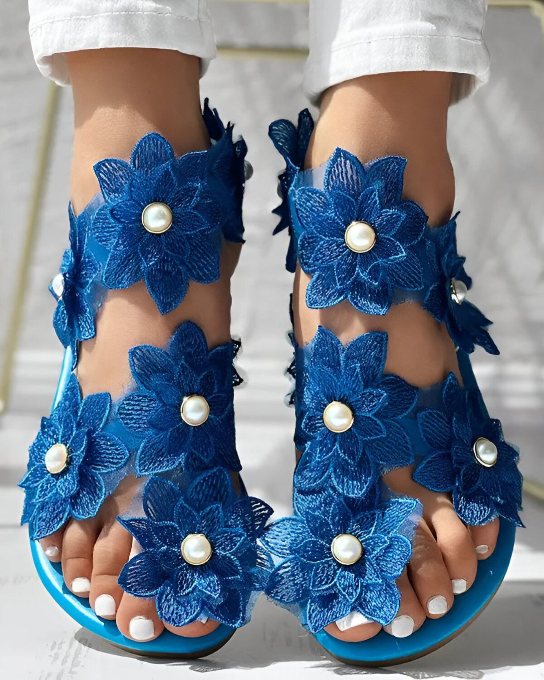 Une paire de sandales plates bleues avec des fleurs en dentelle et des perles. Les sandales sont portées par une femme qui porte un jean blanc et un haut blanc.