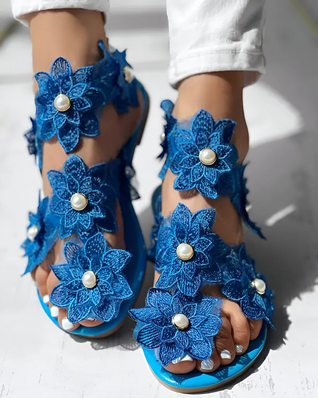 Une femme porte des sandales plates bleues avec des fleurs en dentelle et des perles. Les sandales sont ouvertes au niveau des orteils et du talon. La femme porte un jean blanc et les ongles des pieds peints en blanc.