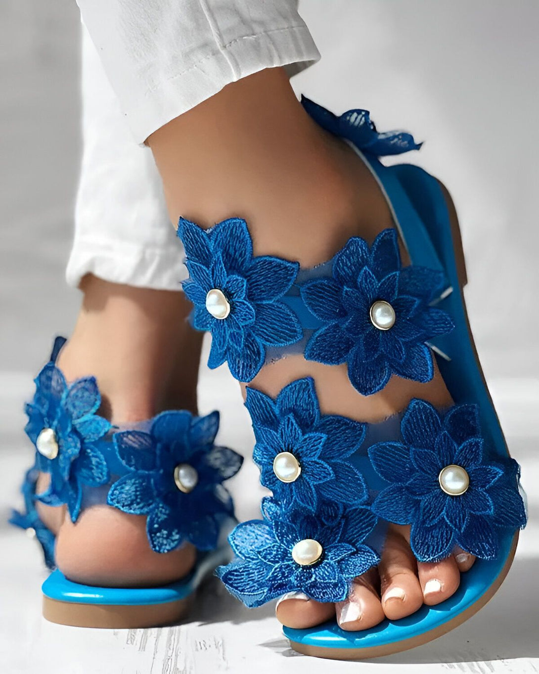 Une femme porte des sandales plates bleues avec des fleurs en dentelle et des perles. Les sandales sont ouvertes au niveau des orteils et ont une bride à la cheville. La femme porte un pantalon blanc et les ongles des pieds peints en bleu.