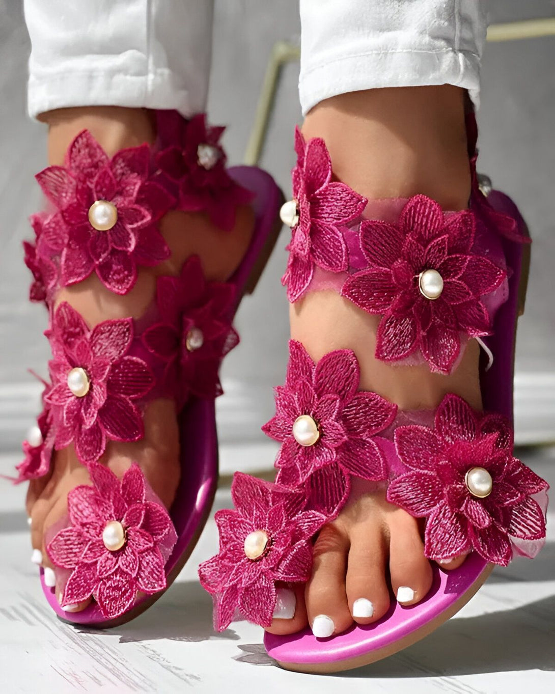 Une femme porte des sandales roses à talons hauts. Les sandales sont ornées de fleurs en tissu et de perles. La femme porte un jean blanc et un haut blanc. Elle est debout sur un sol en bois.