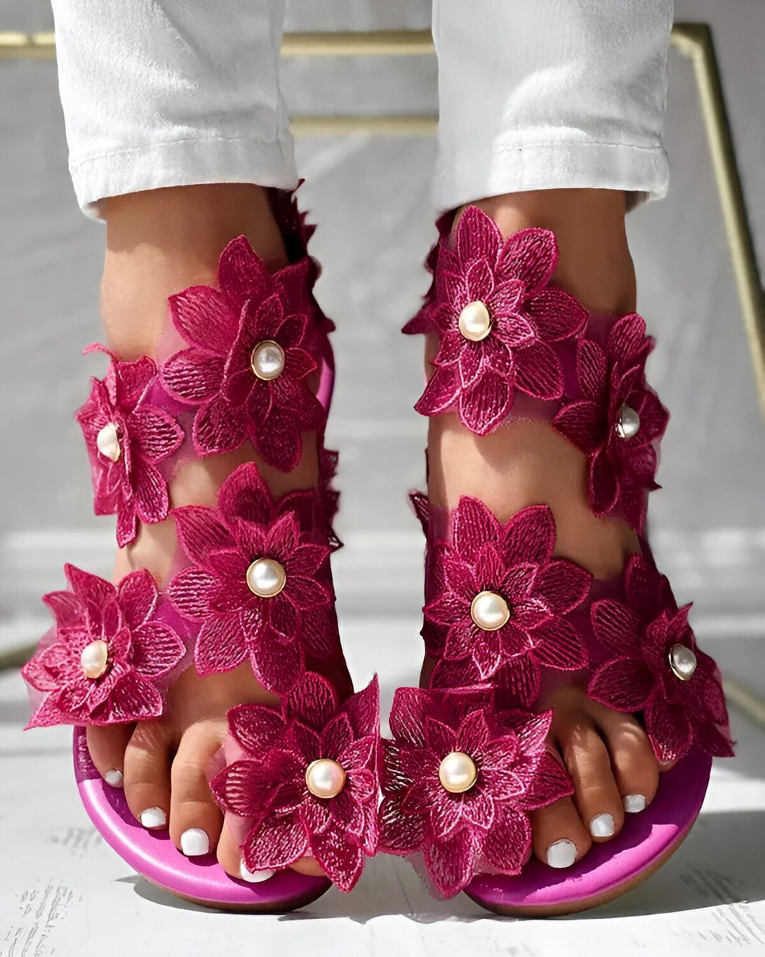 Une femme porte des sandales roses à talons hauts. Les sandales sont ornées de fleurs en tissu et de perles. La femme porte un jean blanc et un haut blanc. Elle est assise sur une chaise et ses pieds sont posés sur le sol.