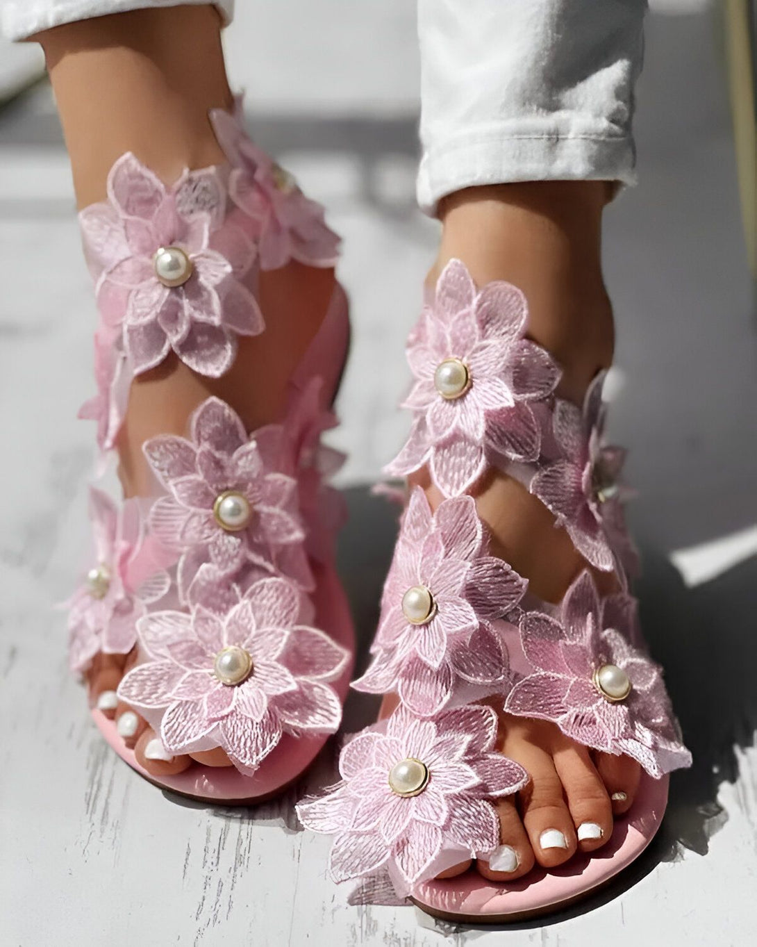 Une paire de sandales roses à talons hauts avec des fleurs en dentelle et des perles. Les sandales sont portées par une femme qui porte un jean blanc et un haut blanc.
