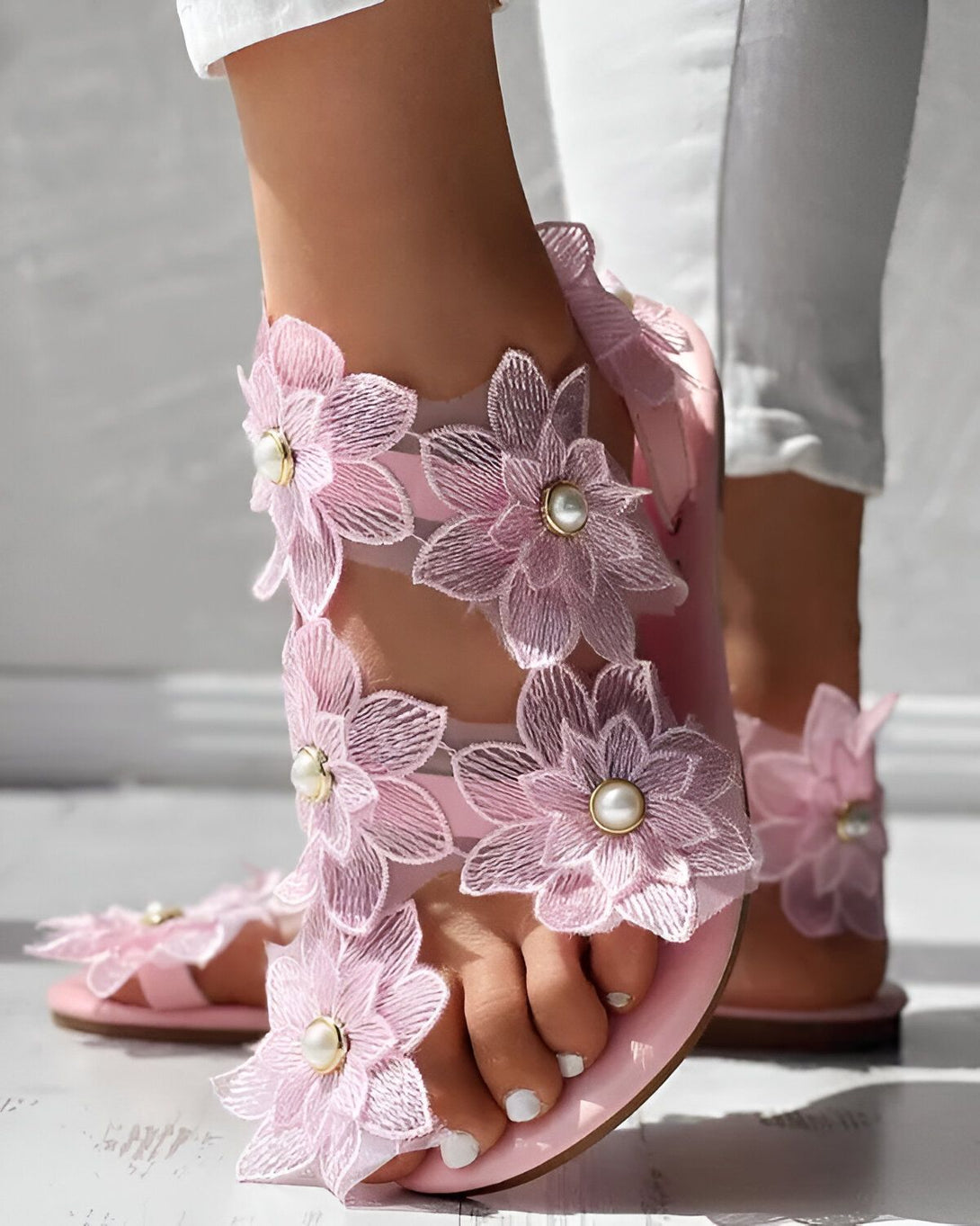 Une femme porte des sandales roses à fleurs. Les fleurs sont en 3D et ont des perles au centre. Les sandales sont plates et ont une bride à la cheville. La femme porte un jean blanc et un haut blanc. Elle est debout sur un sol en béton.