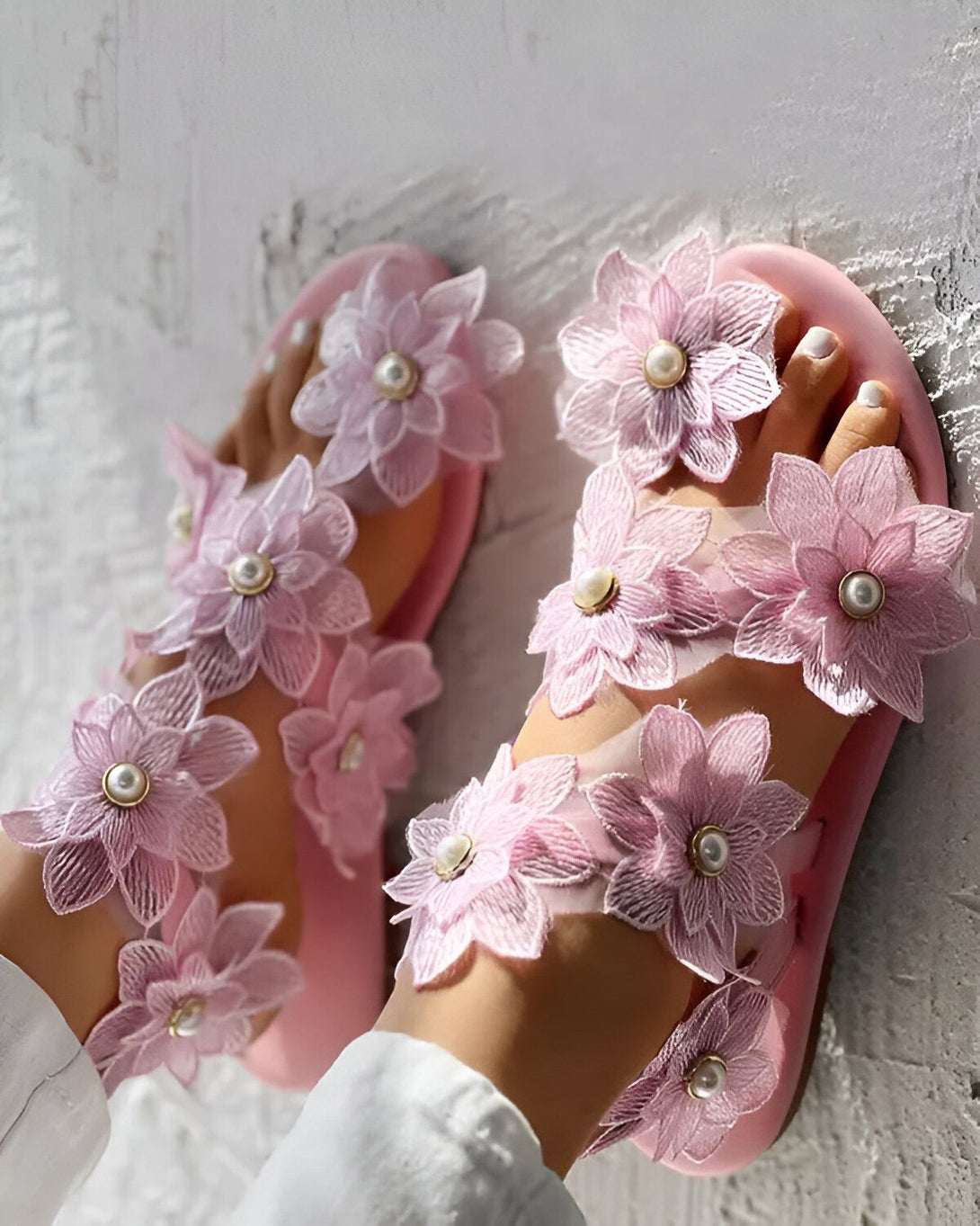 Une paire de sandales roses à talons plats avec des fleurs en tissu et des perles. Les sandales sont portées par une femme qui se tient debout sur un sol en béton.