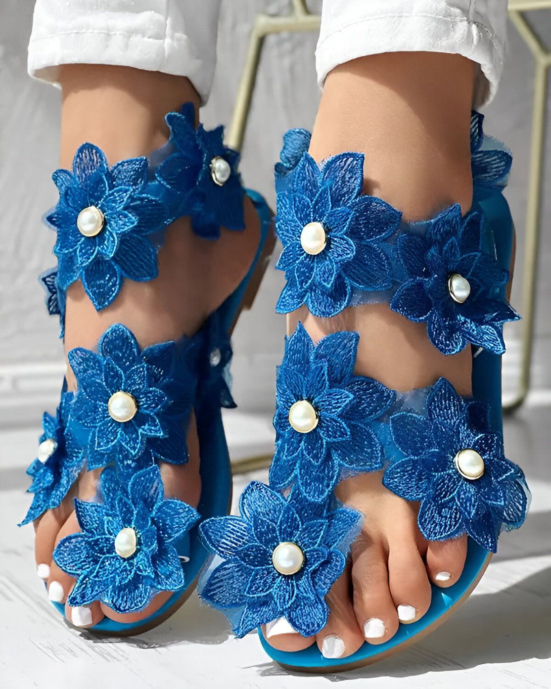 Une paire de sandales plates bleues avec des fleurs en dentelle et des perles. Les sandales sont portées par une femme qui porte un pantalon blanc et un haut blanc. Les sandales sont élégantes et féminines et peuvent être portées pour une variété d'occasions.