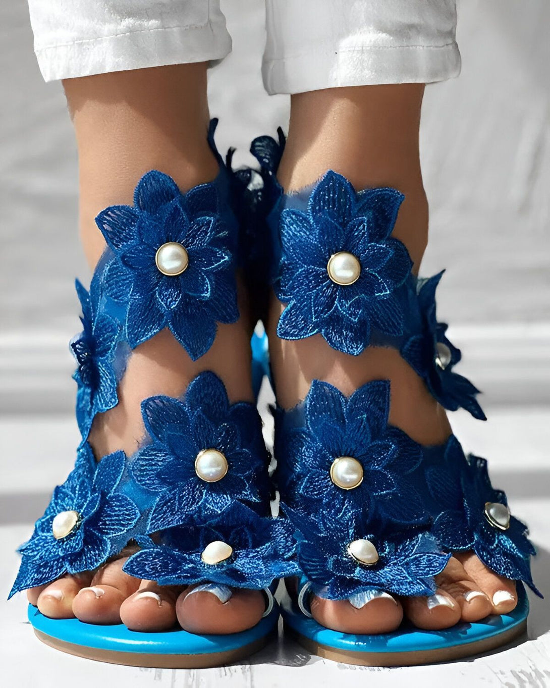 Une paire de sandales plates bleues avec des fleurs en dentelle et des perles. Les sandales sont portées par une femme qui porte un jean blanc et un haut blanc.