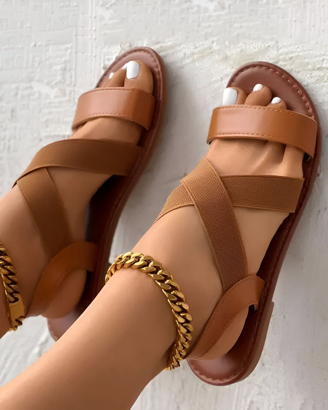 Une femme porte des sandales marron avec des brides croisées sur le dessus du pied. Les sandales sont en cuir et ont une semelle plate. La femme a les ongles des pieds peints en blanc et porte un bracelet de cheville doré.