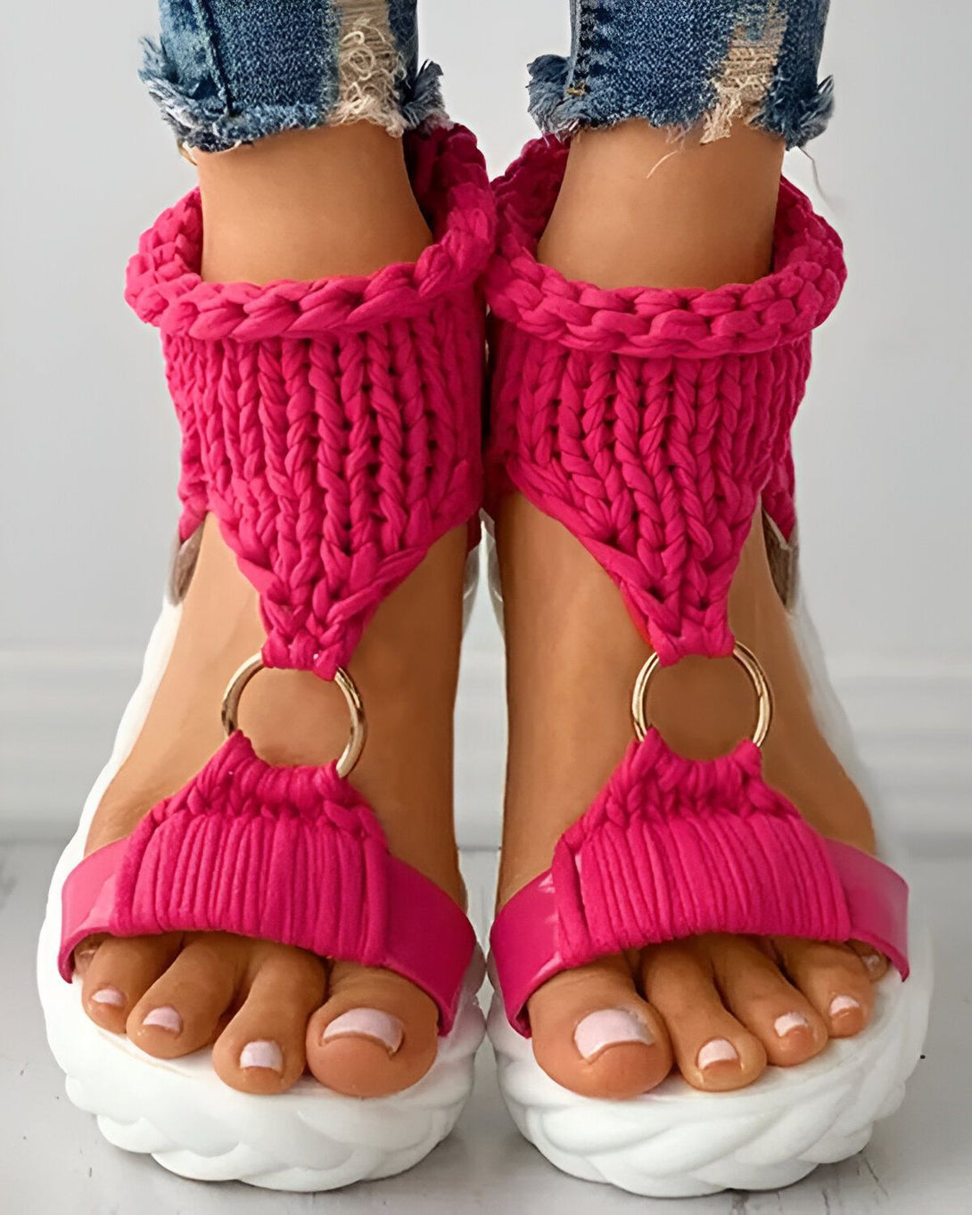 Une femme porte des sandales roses avec des lacets qui s'enroulent autour de la cheville. Les sandales ont une semelle blanche et une bride qui passe entre les orteils. La femme porte un jean bleu et les ongles des pieds peints en rose.