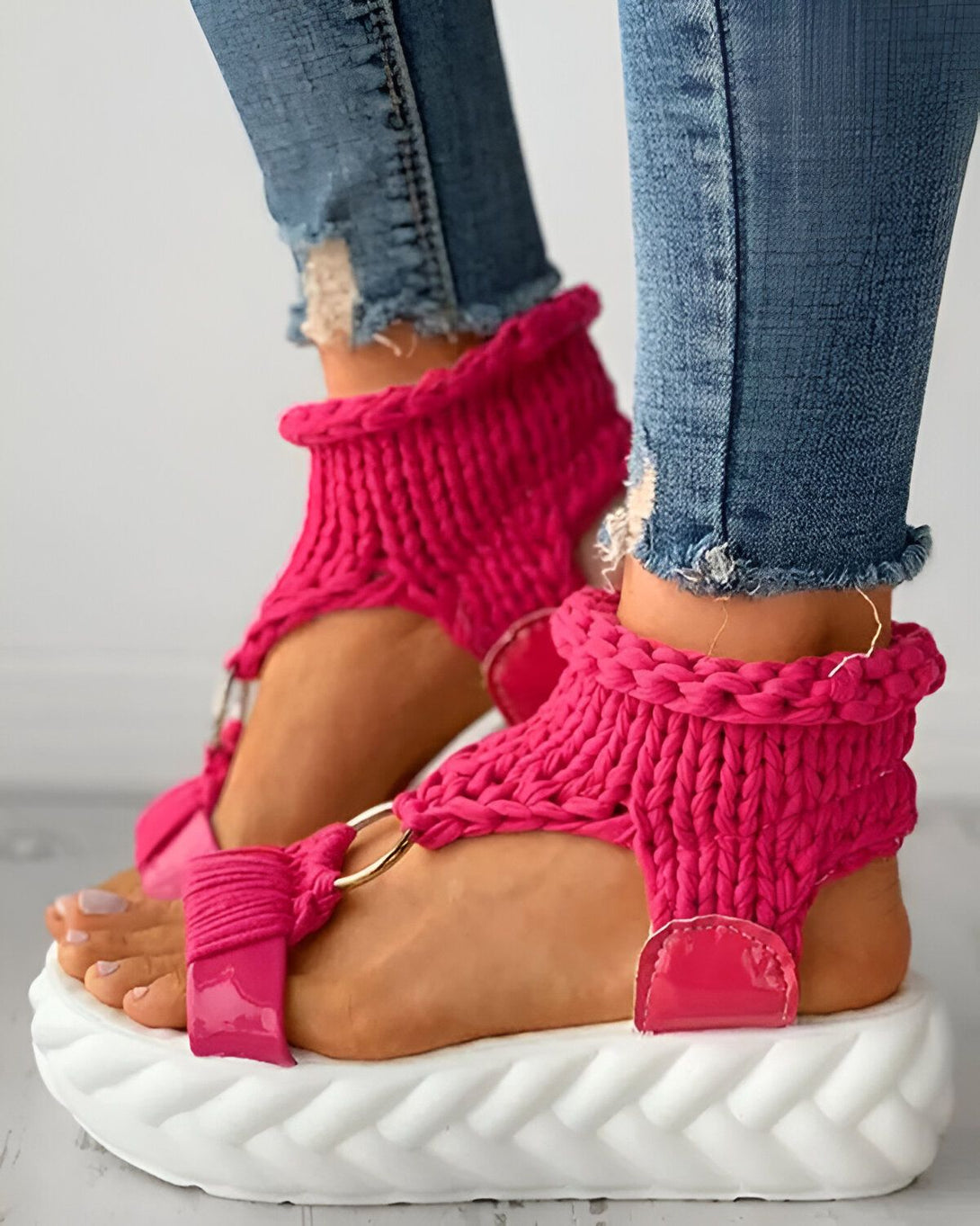 Une femme porte des sandales roses en laine avec un jean bleu troué. Les sandales ont une semelle blanche épaisse et des brides qui s'enroulent autour de la cheville.