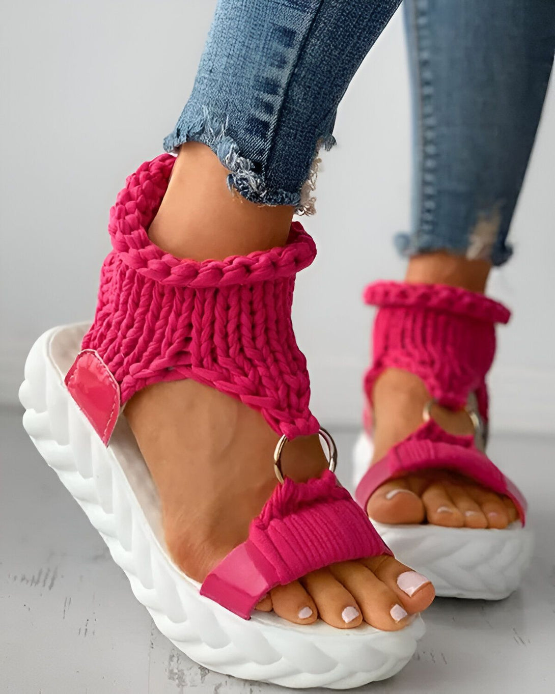 Une femme porte des sandales compensées roses. Les sandales sont en laine et ont une bride qui s'enroule autour de la cheville. La femme porte un jean bleu et les ongles des pieds peints en rose.