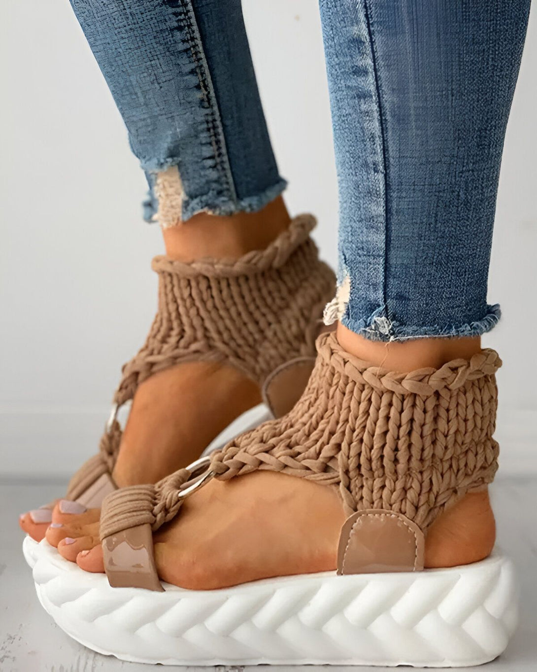 Une femme porte des sandales marron clair avec des semelles compensées blanches. Les sandales sont faites d'un matériau tissé et ont des brides qui s'enroulent autour de la cheville. La femme porte un jean bleu troué.