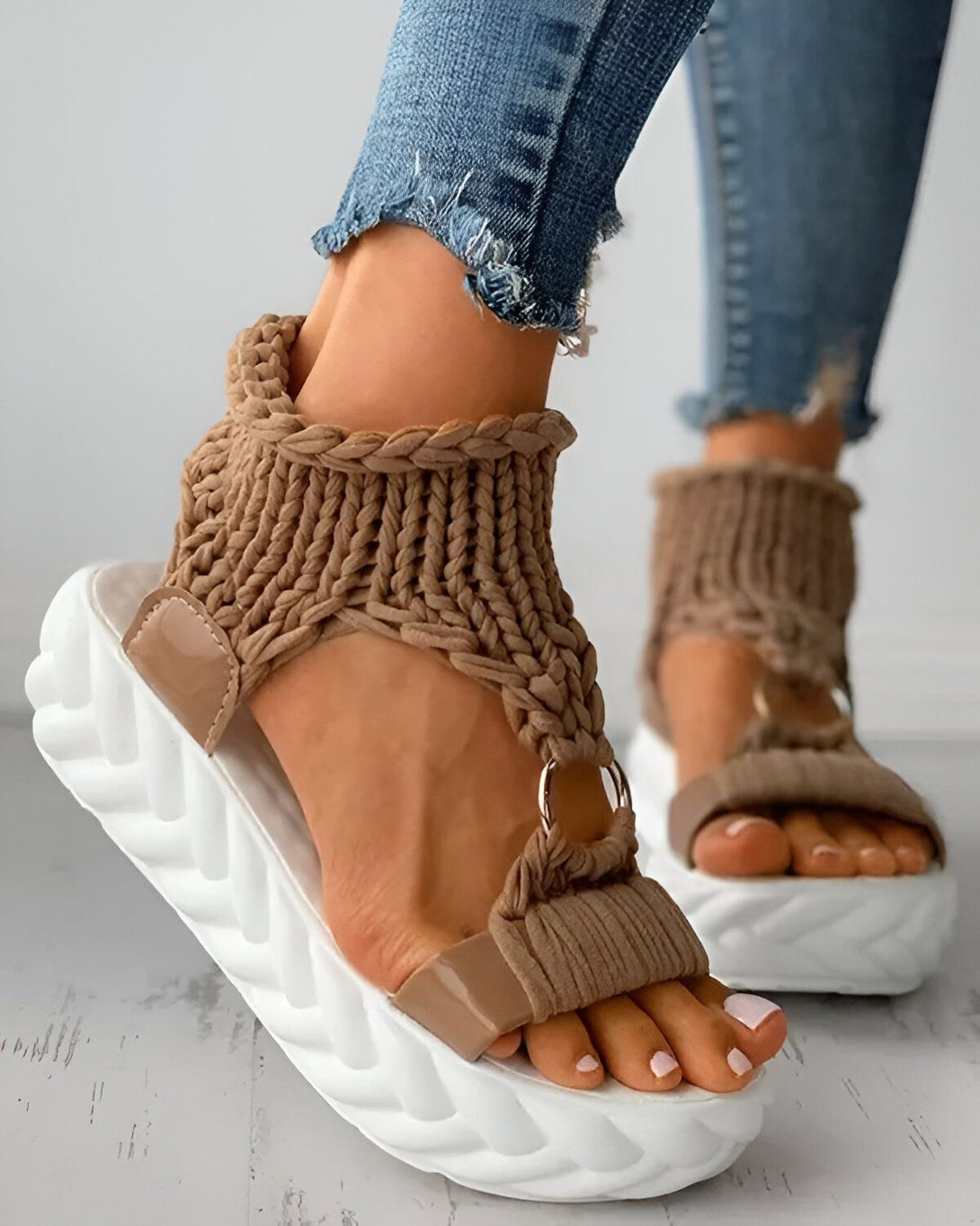 Une femme porte des sandales marron clair avec une semelle blanche. Les sandales sont faites d'un matériau tricoté et ont une bride qui s'enroule autour de la cheville. La femme porte un jean bleu et les ongles des pieds peints en rose.