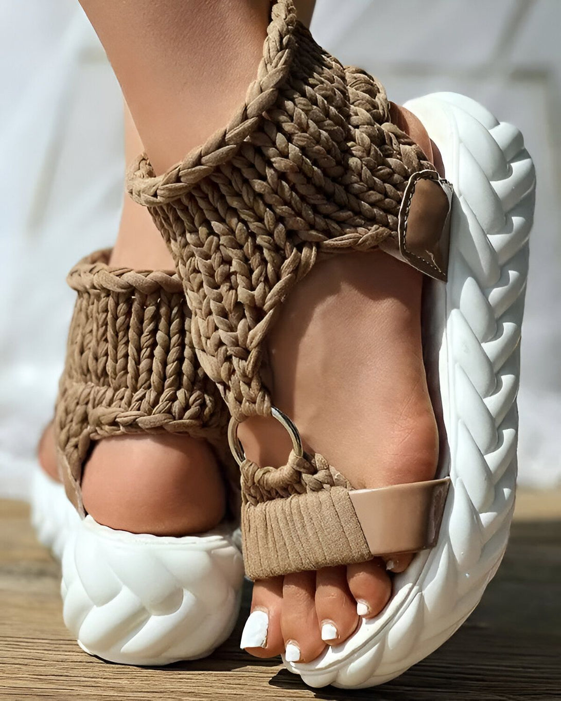 Une femme porte des sandales marron et blanches. Les sandales sont faites de corde tressée et ont une semelle épaisse. La femme a les ongles des pieds peints en blanc.
