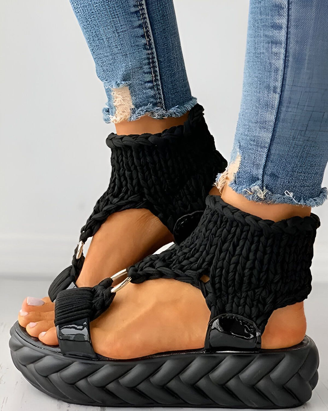 Une femme porte des sandales noires à talons compensés. Les sandales sont en corde tressée et ont des brides qui s'enroulent autour de la cheville. La femme porte un jean bleu déchiré et les ongles des pieds peints en rose.