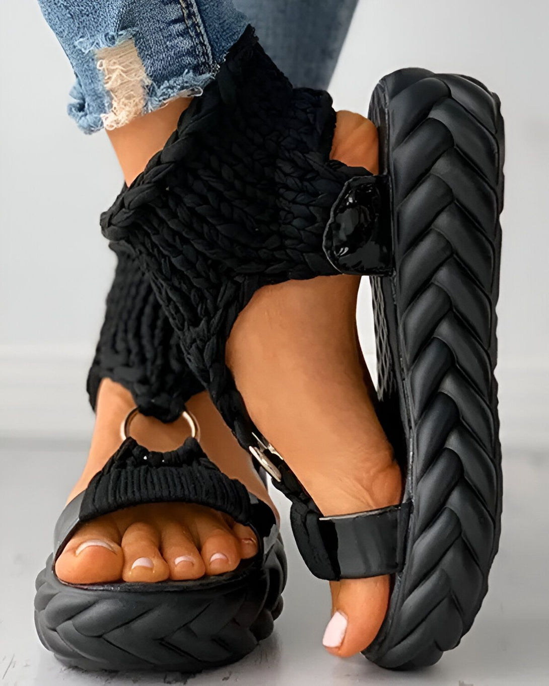 Une femme porte des sandales noires à plateforme. Les sandales sont en matière synthétique et ont une bride qui s'enroule autour de la cheville. La femme porte un jean bleu troué.