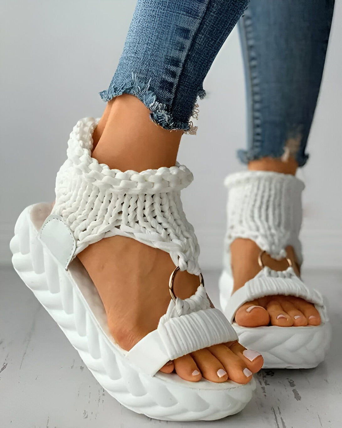 Une femme porte des sandales blanches compensées. Les sandales sont en laine et ont une bride qui s'enroule autour de la cheville. La femme porte un jean bleu et les ongles des pieds peints en rose.