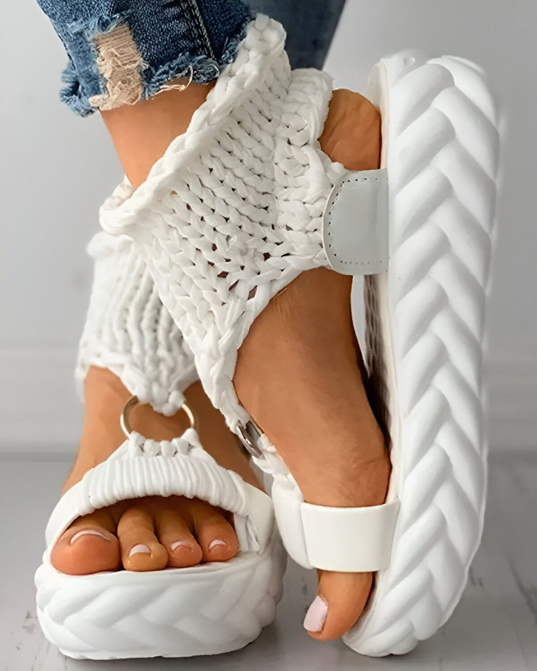 Une femme porte des sandales blanches compensées. Les sandales sont en matière synthétique et ont une bride qui s'enroule autour de la cheville. La femme porte un jean bleu troué.