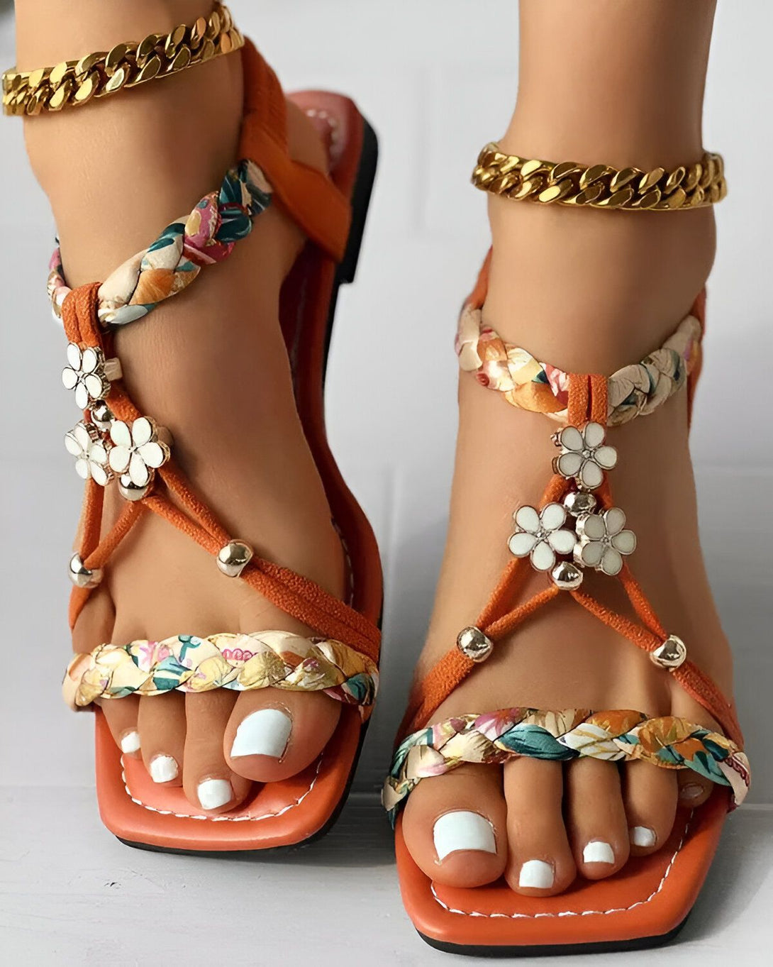 Une paire de sandales plates orange à brides croisées sur le dessus du pied. Les brides sont décorées de fleurs blanches et de feuilles vertes. Les sandales sont portées par une femme qui a les ongles des pieds peints en blanc.