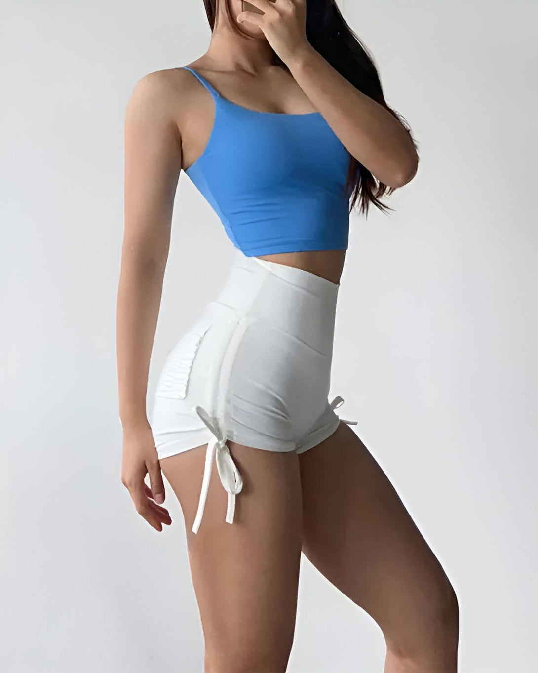 Une femme porte un soutien-gorge de sport bleu et un short blanc. Elle est vue de côté et tient ses cheveux longs et noirs avec sa main droite.