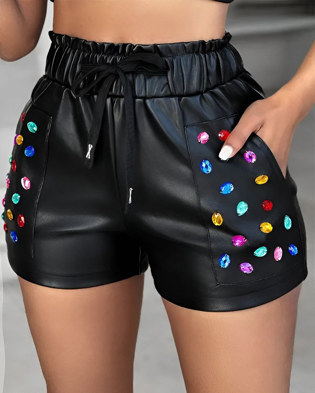Une femme porte un short en cuir noir avec des pierres précieuses colorées sur la poche droite. Le short est taille haute et possède un cordon de serrage à la taille.