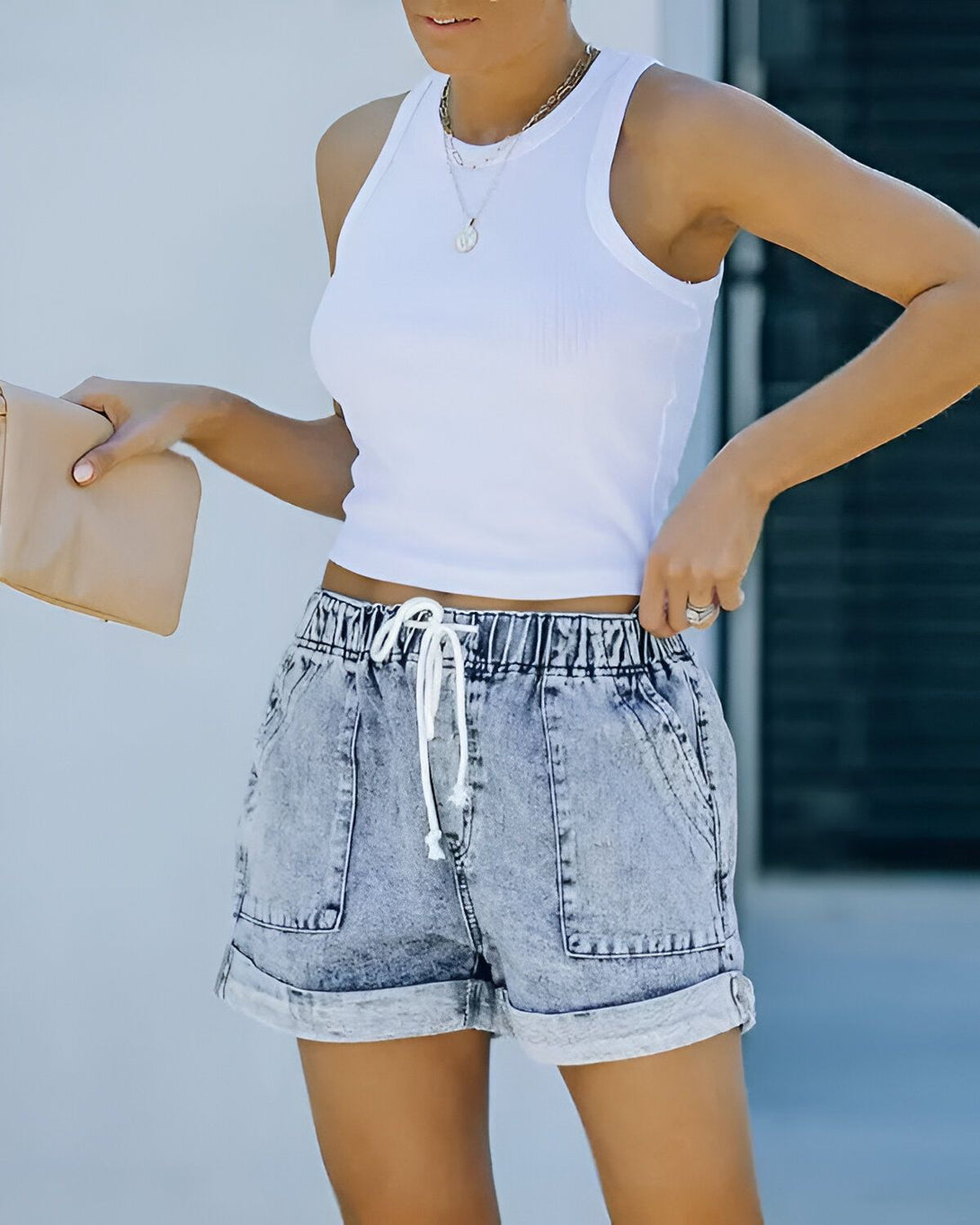 Une jeune femme porte un débardeur blanc et un short en jean bleu. Elle a un sac à main beige à la main.