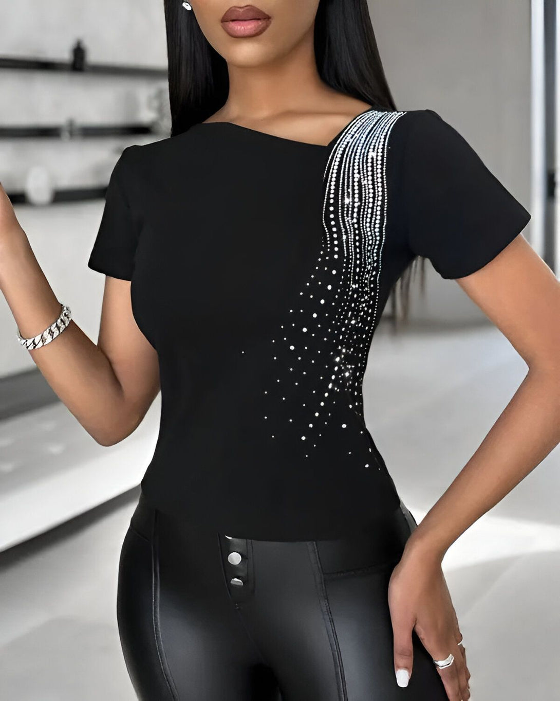 Une femme porte un chemisier noir à manches courtes avec des strass sur l'épaule. Elle porte également un pantalon en cuir noir et des talons noirs.