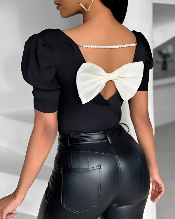 Une femme porte un chemisier noir avec un grand nœud blanc dans le dos. Le chemisier est à manches bouffantes et le nœud est noué avec une chaîne de perles. La femme porte également un pantalon en cuir noir.