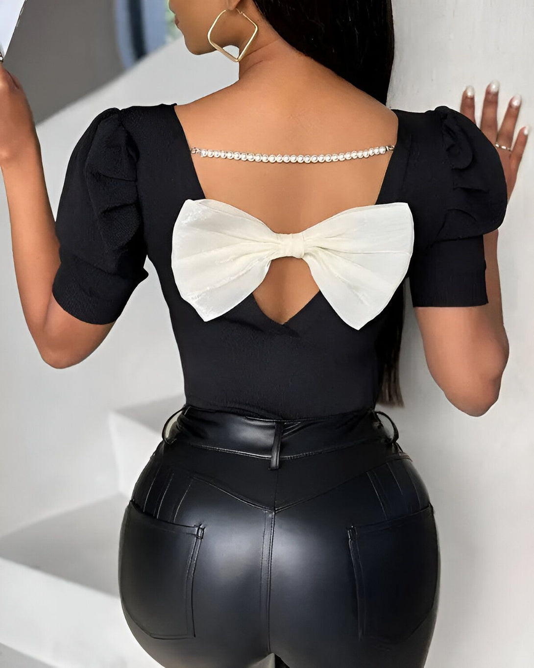 Une femme porte un chemisier noir avec un grand nœud blanc dans le dos. Le chemisier est en dentelle et a des manches bouffantes. La femme porte également un pantalon en cuir noir.