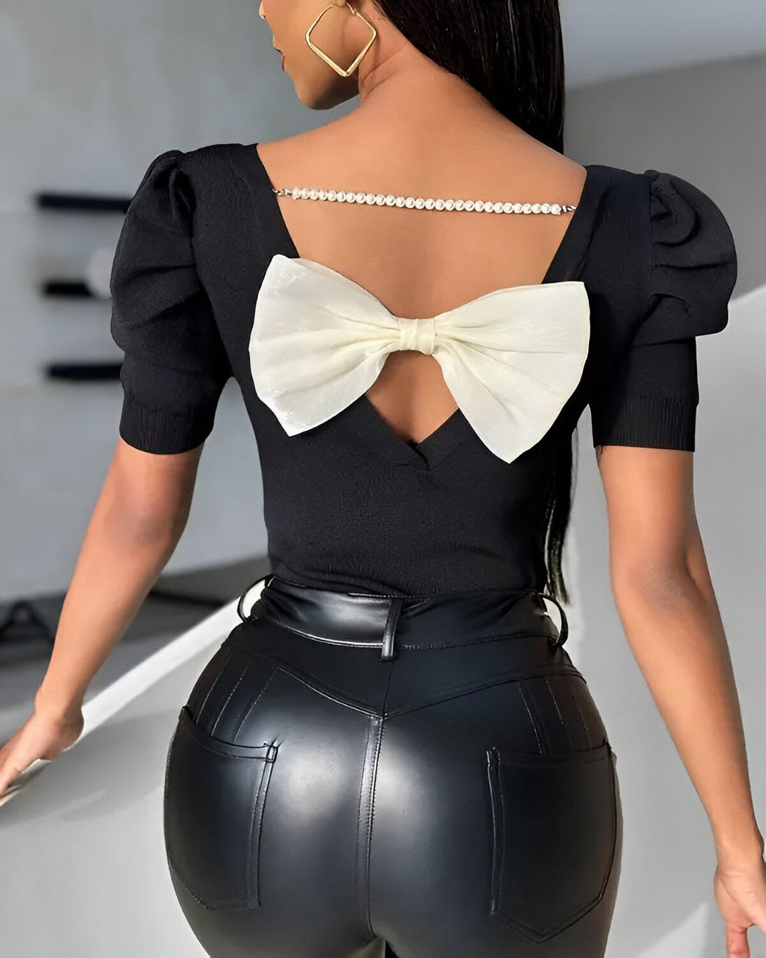 Une femme porte un chemisier noir avec un grand nœud blanc dans le dos. Le chemisier est à manches courtes et a un col montant. La femme porte également un pantalon en cuir noir.