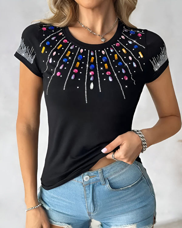 Une femme porte un t-shirt noir avec des strass multicolores. Le t-shirt est moulant et a des manches courtes. La femme est blonde et a les yeux bleus. Elle porte un jean bleu et des talons noirs.